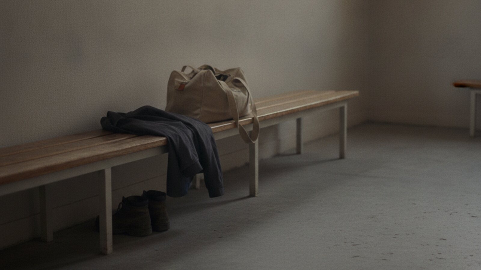 Quiet changing-room bench in early morning light with a folded dark jacket, a canvas kit bag and boots beneath it at the end of a shift